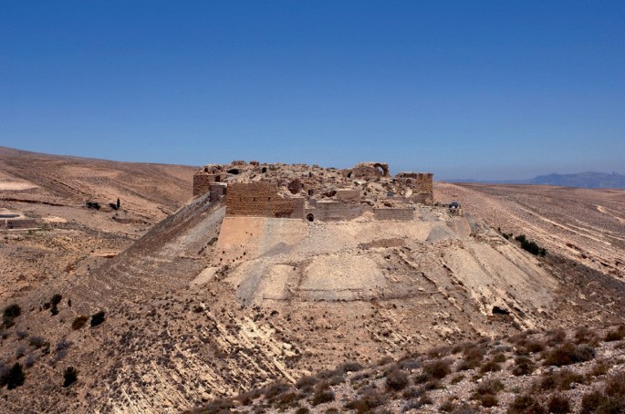 A photograph of a large, rocky hill with a small castle on top. The castle is made of stone and has several windows and doors. The hill is surrounded by a barren landscape with no vegetation or trees. The sky is clear and blue.