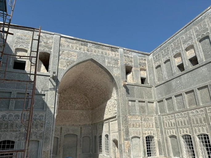 A photo presents the bottom-angle view of a large multi-storied building. It has ornate walls and a prominent archway. A scaffolding appears in partial view on the left and some of the windows and doors are missing.