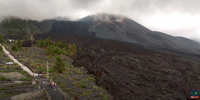 A photograph of a volcano with smoke emitting from it. There is vegetation with trees, bushes, and a road with towers on the left. There are people on the road.