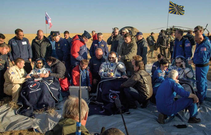 A photograph displays 3 astronauts who returned from the International Space Station. Many other support staff and teammates are there.
