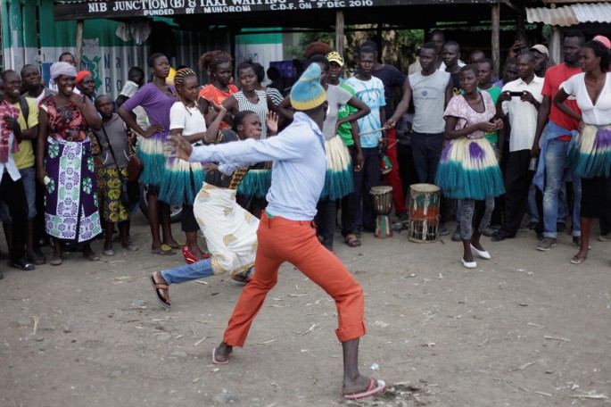 A photo of a group of people performing in an open public space. A man stands facing away from the camera and swings his right arm wide backwards as a woman slightly crouches, putting her hands in front as if to defend a strike. A crowd watches them perform in the background.