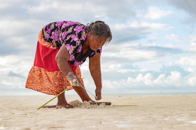 A photograph of a woman collecting sea worms at the coastline under a cloudy and sunny sky.