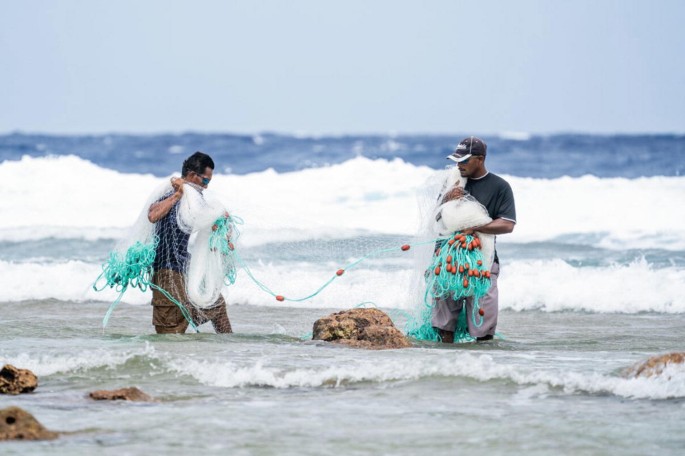 A photograph of two fishermen stands in the sea, holding a fish net, against the backdrop of rolling sea waves.