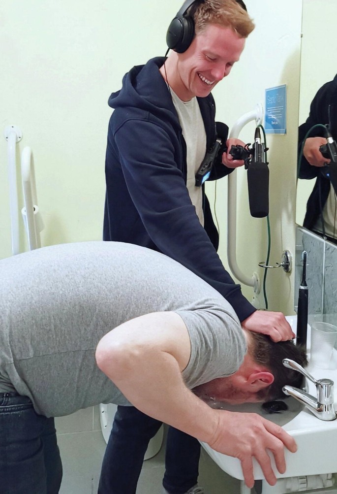 A photograph of a man wearing headphones and holding a large microphone, pressing the head of another man into a wash basin filled with water.