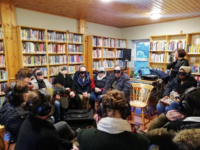 A photograph of a group of people who have their eyes covered, seated inside a library and listening to their headphones.