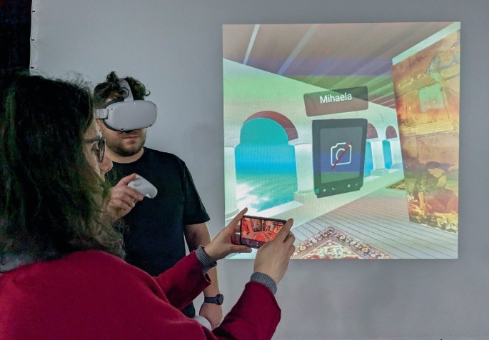 A photograph of 2 people in a V R room. One person wears a V R headset and holds 2 controllers in their hand. Another person holds a smartphone with the V R enviroment on screen. The wall behind has a projection of the V R environment of Villa Storck.