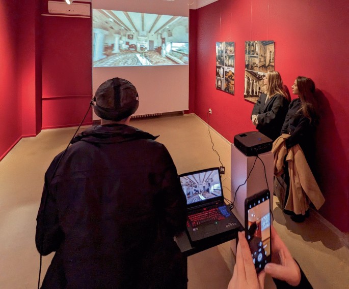 A photograph of a V R room with a projection of a V R environment on a screen. A person wears a V R headset connected to a laptop and faces the projection. 2 people stand near a wall with museum displays. Another person captures the room on their smartphone.