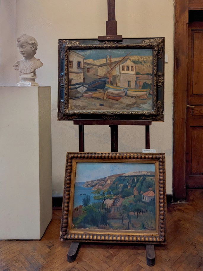 A photograph of 2 framed paintings and a bust in a museum with a wooden floor. One painting is mounted on an easel, while another is placed on the easel's base. The bust is placed on a column near the easel. A wooden door is in the background.