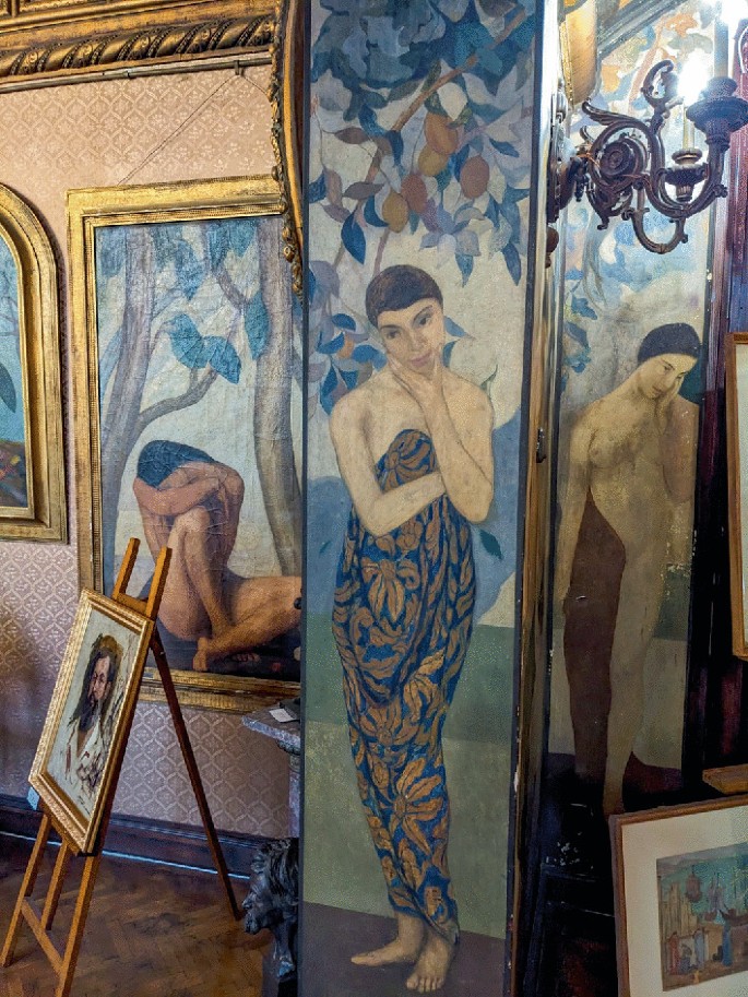 A photograph of tall painted panels mounted on gilded walls of a museum. The panels depict women in different poses. Smaller framed paintings on easels are kept on the wooden floor.