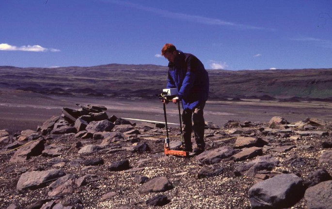 A photograph of an archaeologist collecting earth resistance data on a hilly, stony area of Steinastadir, Iceland.