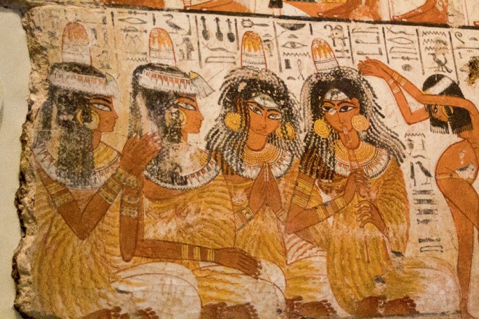A painting of 4 women dressed in traditional attire with headdresses, earrings, bangles, and necklaces. One woman on the left claps her hands, another in the middle also claps, and the third woman holds two musical pipes in her mouth. On the far right, a naked woman turns her upper body while clapping her hands.