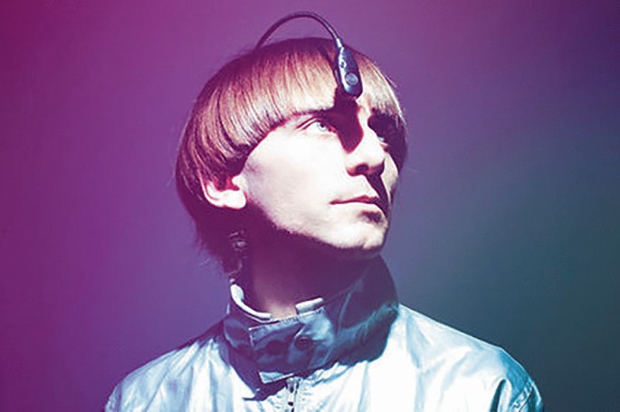 A photo of Neil Harbisson who wears an eyeborg that extends outward from the back of his head. The frame containing the antenna rests against his forehead.