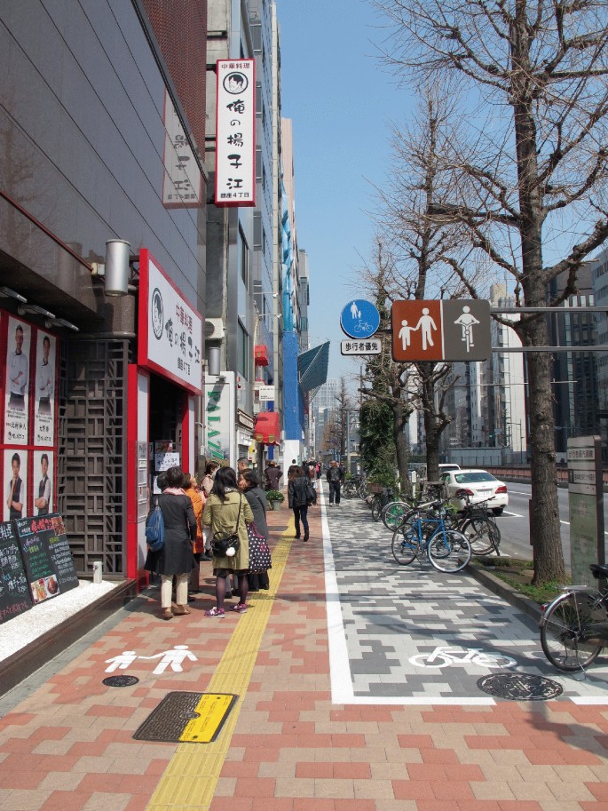 A photo of a pathway lined with signboards for walking and cycling. On one side, trees border the pathway, while the other side is occupied by shops. Groups of people walk along the pathway, and bicycles are parked along the other side.