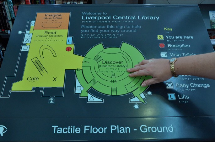 A photo of a tactile floor plan layout of the ground floor. A hand points to the children's library located at the center. To the left of the children's library are areas designated for music and fun, a reading area, and a cafe. The symbols on the plan are mentioned on the right including lifts.