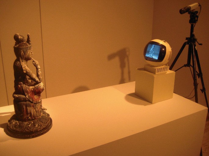 A photo of a dull-lit room where a Buddha statue is placed on the table against a monitor. The monitor is connected to a camera on a tripod behind the monitor. The Buddha statue is reflected on the screen.