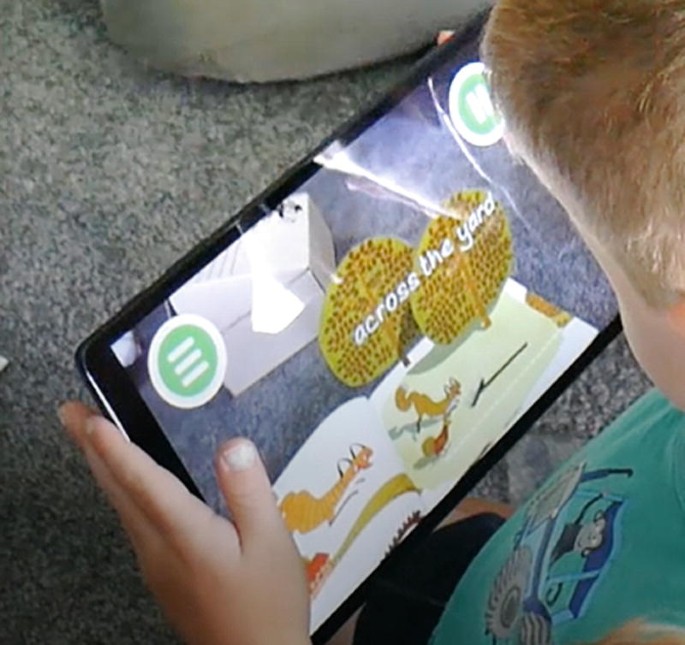 A photo of a young boy using the Rosie app on a tablet. On the screen, the term, across the yard is written across 2 trees and a few animals run across a flat space.