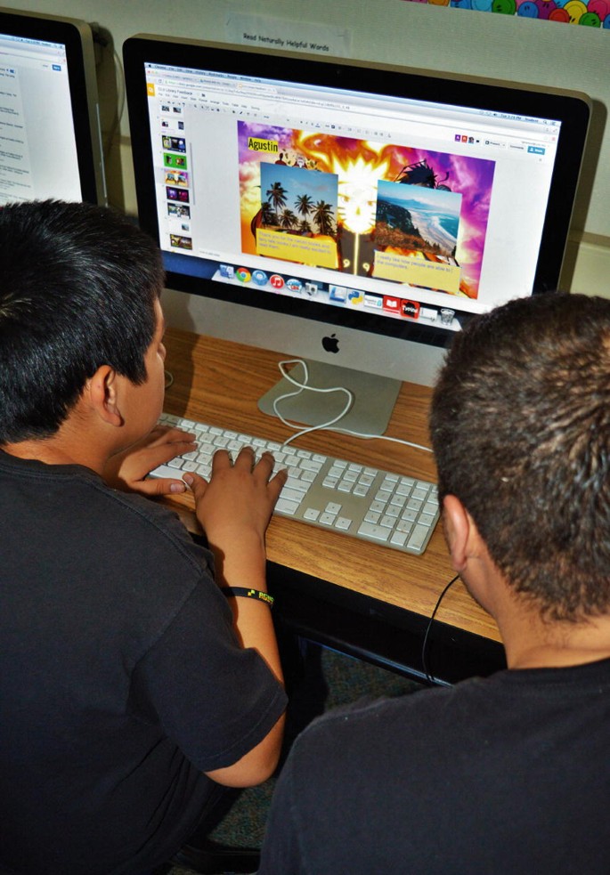 A top-view photo of 2 men who work on an Apple desktop computer. One of the men has his hands on the keyboard. The screen displays the digital artwork with 2 photos and texts.