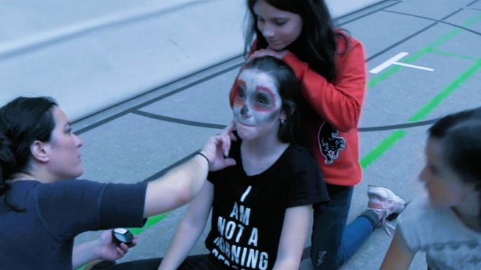 A photo of 2 girls who apply zombie make-up to another girl while sitting on the floor. Another girl gazes while sitting beside them.