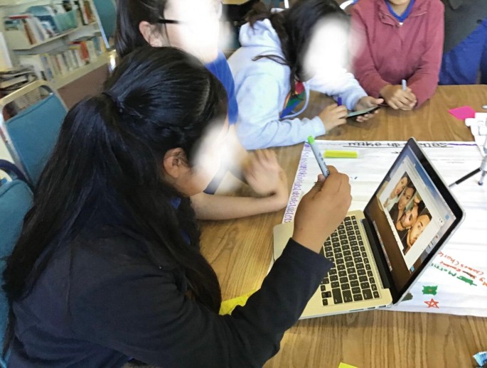 A photo of a group of students gathered around a laptop on a desk. One student appears to be interacting. There are papers and writing materials on the desk.