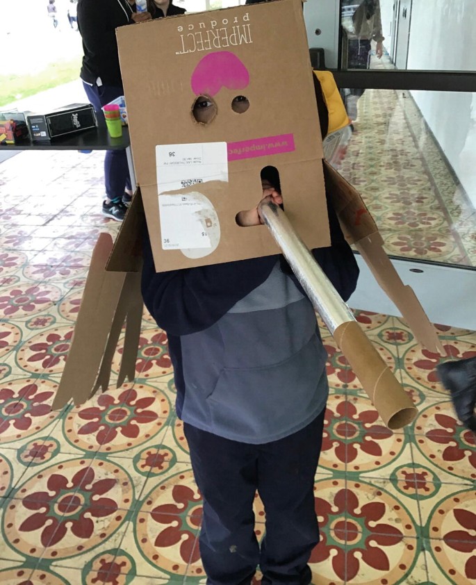 A photo of a standing child who wears a cardboard-box-turned-mask over their head, while holding a long paper tube sticking out from a hole in the box. The box has the text written over it that reads, Perfect produce.