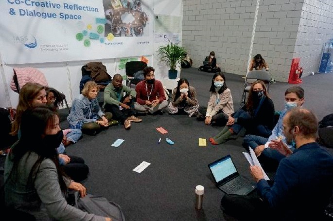 A photo of a group of seated people on the floor. 1 of the people is in mid-speech in front of a laptop, while others gaze at him. A banner with text is on the wall behind them, with a potted plant on a nearby stool. 3 other people sit behind them in a lobby area, lining the wall in the background.