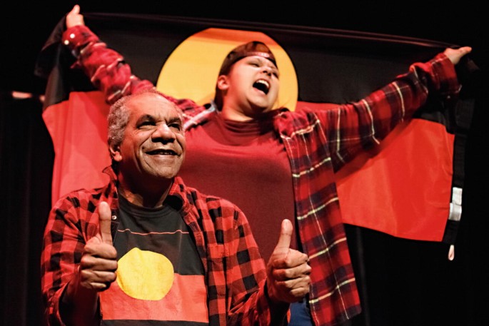 A photo of 2 people on stage. Gregory J Fryer smiles with both hands making a thumbs-up gesture while Nazaree Dickerson stands behind holding a banner with a bright band in the middle and dark bands at the top and bottom. Gregory wears a t-shirt with the same pattern.