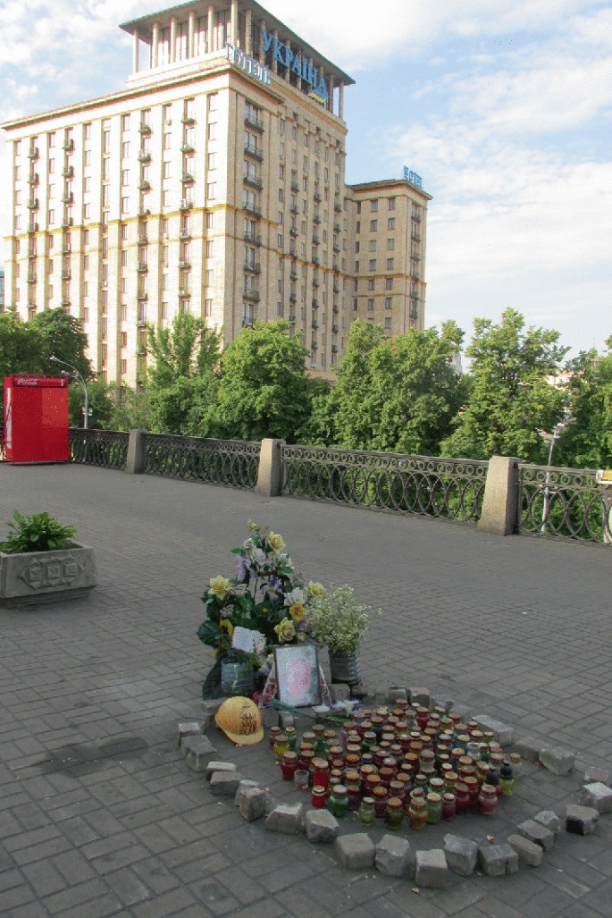 A photo of a makeshift memorial on a paved path with a large multistoried building in the background. The memorial has a few flowers, bouquet, and a photograph placed behind a few stones placed around several jars.