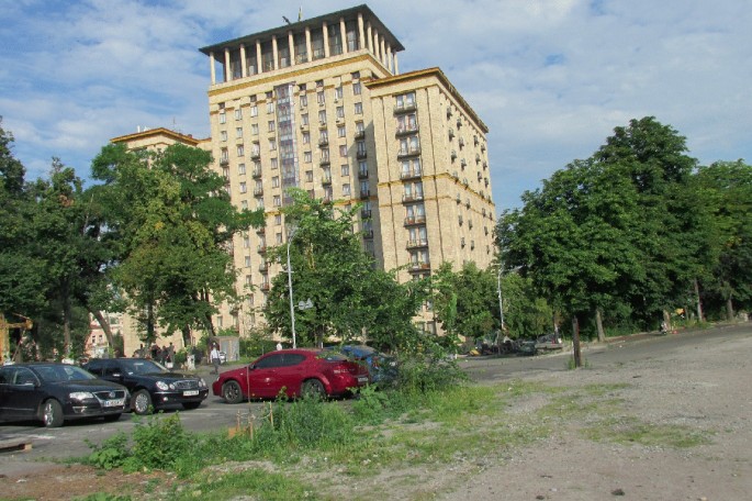 A photo presents a low-angle shot of Hotel Ukraina. It is a large multistoried building with tree frontage and a few cars parked by the side of a tarred road.