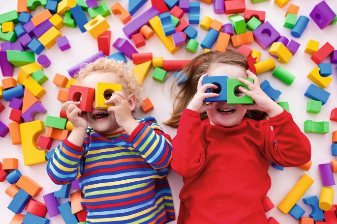 A photo captures two kids playing with various colorful toy blocks. They cover their eyes with the cubical blocks and peep through holes inside them.