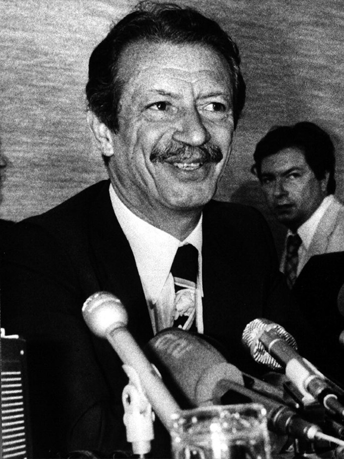 A grayscale portrait photograph of Prime Minister Shapour Bakhtiar in front of microphones.