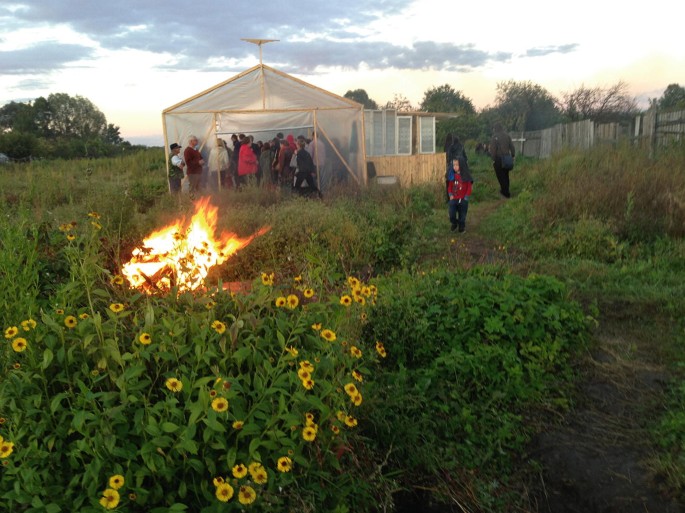 A photograph presenting a small garden culture. A part of the plants has caught fire. There is a tent with most of the people inside and a few people outside it.