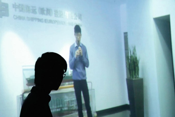 A photograph in which Henry watches footage of himself playing the flute in the entrance hall of the Chinese shipping company.