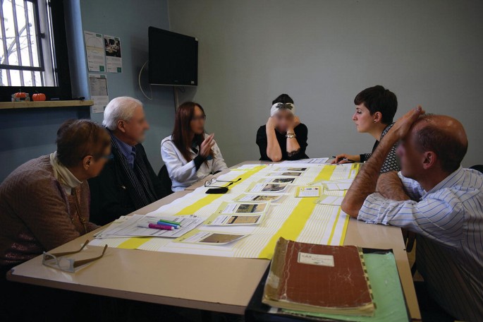 A front view of a group of men and women seated at a table in the middle of a discussion. Several photographs, papers, markers, and a notebook are on the table.