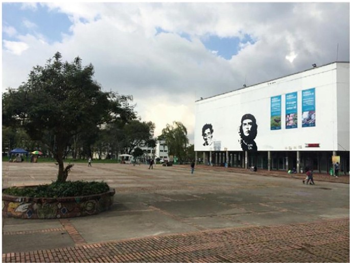 A photo presents a long shot of a large, paved area with a few trees and an open ground. On the left a pavilion appears with a billboard on top. It has a few posters and sketches including that of Che Gue vera's.