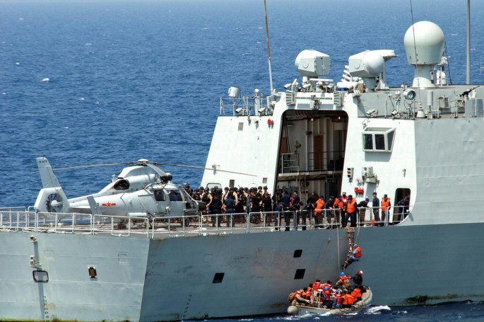 A photograph illustrates people in life jackets boarding a large ship from a small ship in the sea.