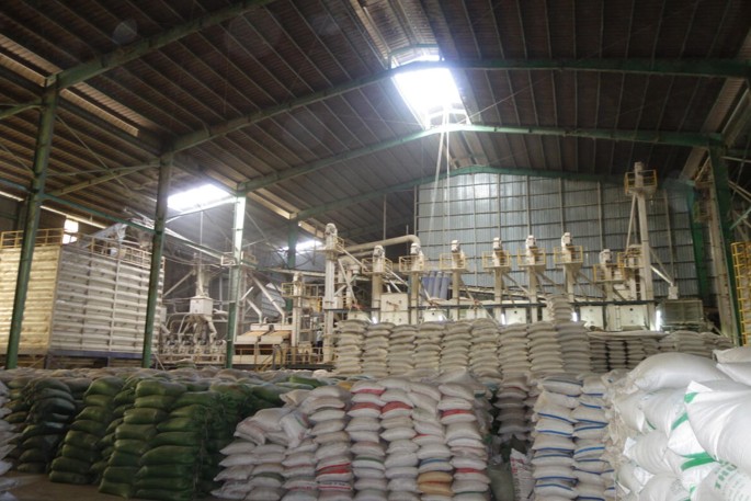 A photograph of the interior of a mill with a shed on top has big machines with pipes and rice sacks stacked on top of one another.