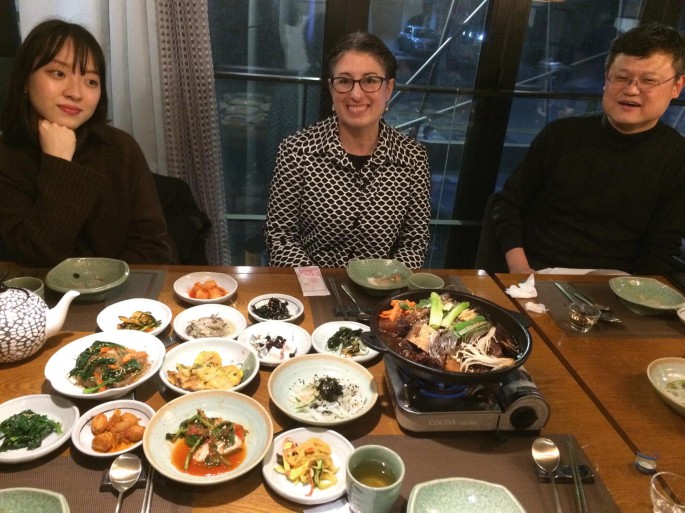 A photograph of two women and a man who are seated at a dining table served with several plates and bowls of food. The woman in the middle smiles at the camera.