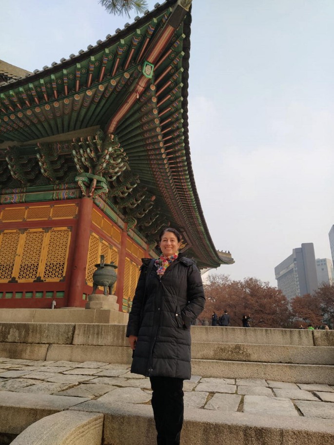 A portrait of a woman standing in front of a monument in Seoul. The monument has a giant hat-like roof. The background has trees and tall multi-storied buildings.