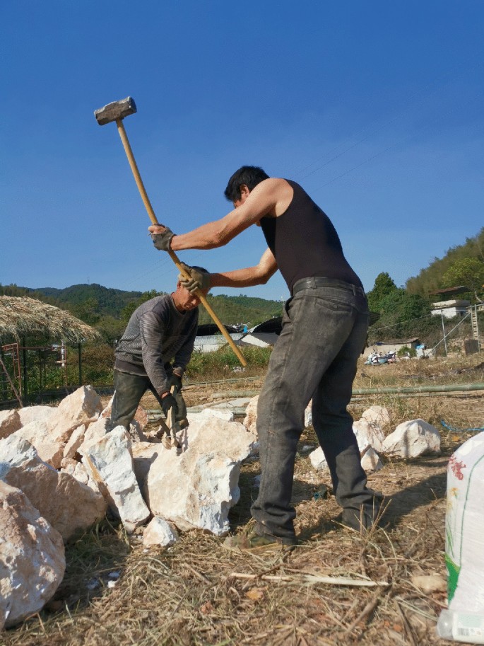A photograph of two men breaking quartz with a hammer. The reconstruction of the shaft furnace is done with the help of stones such as sand, clay, and charcoal powder.