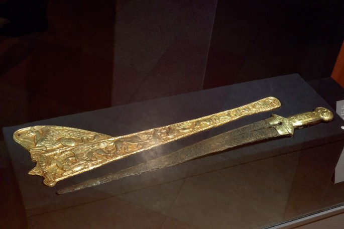 A photograph of a brown sword with a golden hilt and a golden sheet beside it. It is popularly called akinaka and is popularly used in the Persian army at that time.