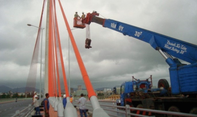 A photograph of field testing on Tran Thi Ly stay cable bridge. Two workers stand on the crane as they install accelerometers for a damping test.