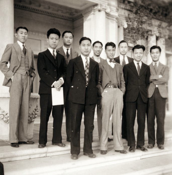 An image of a group of nine people standing in front of building premises, all wearing formal dress.