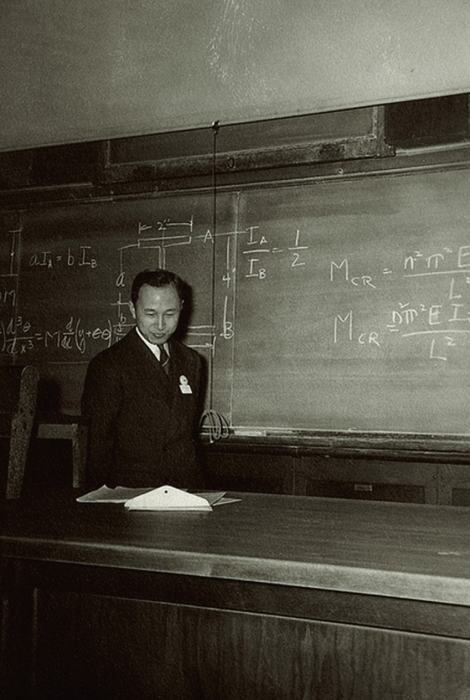 A photograph of a lecturer standing in front of a blackboard with some text written on it.
