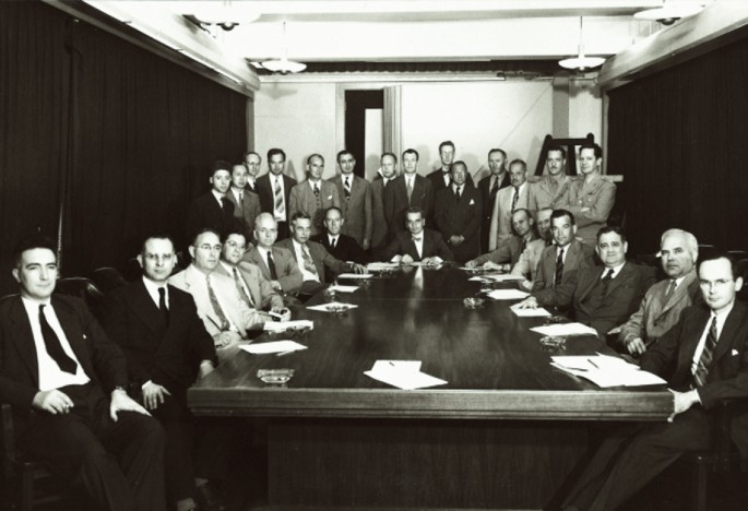 A photograph of the conference room, which includes twenty-eight people. A total of fourteen people, sitting and the same number standing are posing for the camera.
