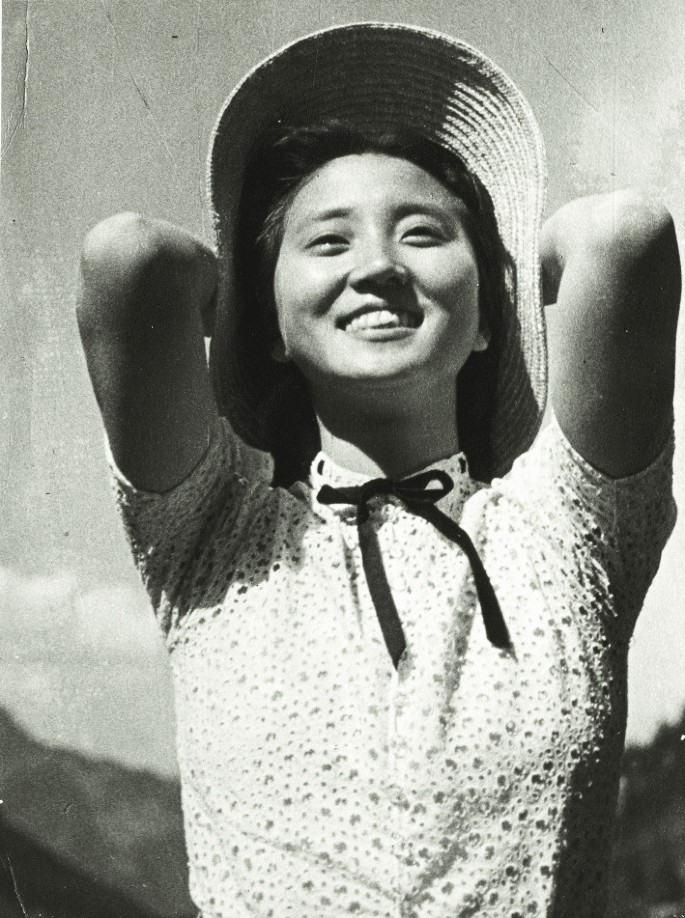 A black-and-white photo of Jiang Ying in her youth. Posing for a photo while grinning and holding a hat with both hands on the head.
