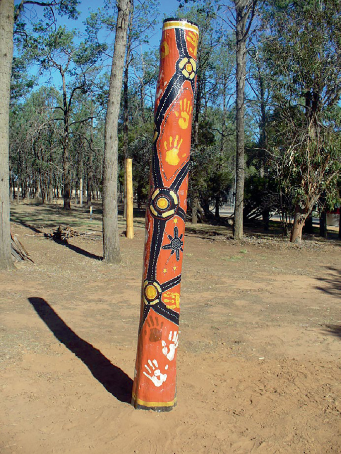 A photograph of a designed and painted totem. The background has dense trees.
