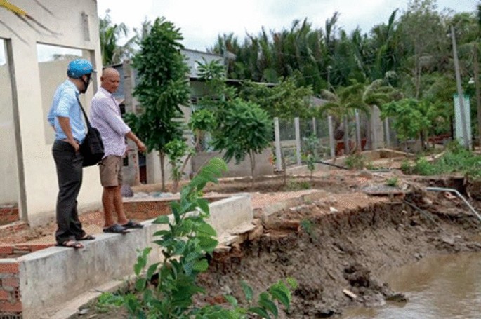 A photo displays 2 men standing on the boundary wall and inspecting the erosion of the road. The muddy water is below the wall.