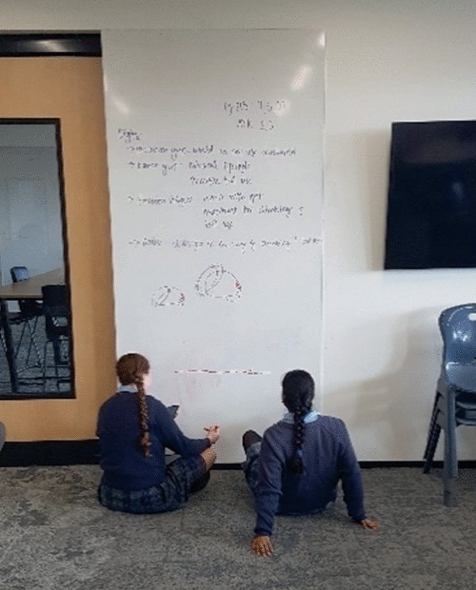 A photograph of a classroom with a large ceiling to floor white board mounted on a wall near the door. Two students are seated on the floor, before the board. A near the board. A math problem has been written on the board. Beside the white board, on the right is a partial view of a large flat screen T V.