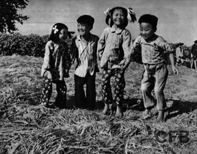 A photograph of 4 children jumping on a heap of thick stalks placed in a field. People in hats, and trees are in the background.