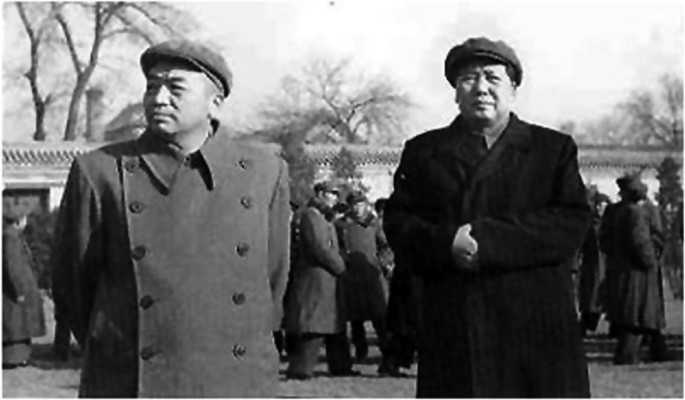 A photograph of Mao Zedong on the right and another man on the left, in overcoats and caps. People in overcoats, buildings, and trees are in the background.